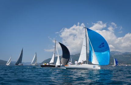 A vibrant array of sailing boats, representing various classes, engage in exhilarating races within the scenic confines of Tivat Bay. Many vessels effortlessly glide downwind, propelled by the natural forces of nature.