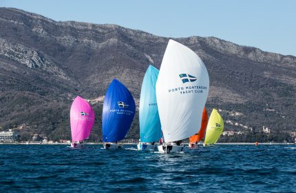 Six vibrant sailing boats form a dynamic pyramid formation as they race towards the shore with colourful sails billowing in the wind.